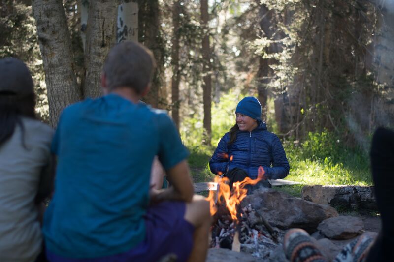 A group of people are gathered around a campfire in a forest setting. The fire is burning brightly, casting a warm glow on the faces of those nearby. One person, wearing a blue jacket and hat, is smiling and looking towards the fire. The others are partially visible, suggesting a close-knit group enjoying the warmth and ambiance of the campfire in a natural environment.
