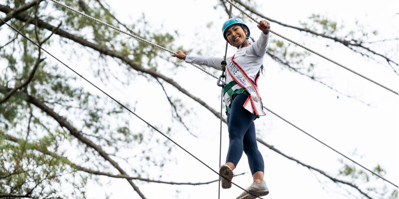 A young girl wearing a helmet and a sash is carefully walking on a tightrope suspended between trees. She is smiling and appears to be enjoying the adventure, with her arms outstretched for balance. The activity seems to be part of an outdoor recreational program or a challenge.