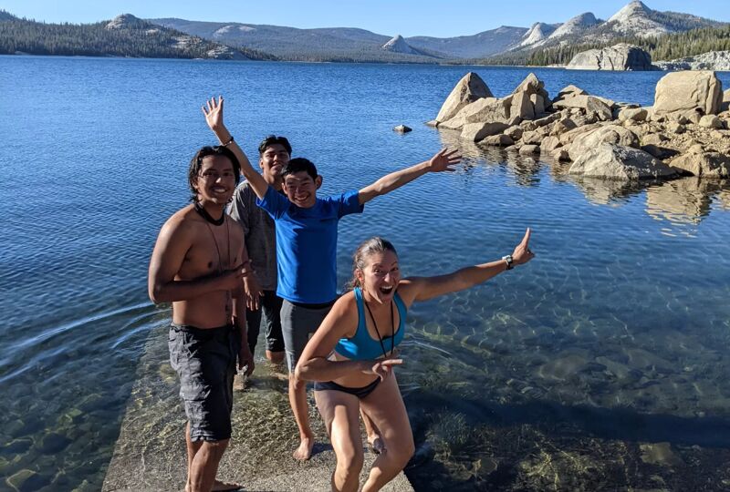 The image shows a group of five young adults standing on a concrete structure that extends into a clear lake. They are all smiling and posing with their arms outstretched in a celebratory manner. The background features a scenic view of mountains and large rocks, suggesting a natural, possibly recreational, setting. The overall atmosphere of the image is joyful and adventurous.