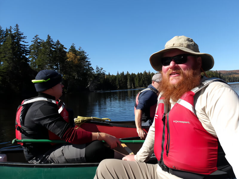 A sunny day on the water shows three people in a canoe. The person in the front is wearing a black hat and a black jacket with a red life vest. The person in the middle is partially visible, wearing a blue shirt. The person in the back is wearing a tan hat and a red life vest. They all appear to be enjoying a leisurely paddle.