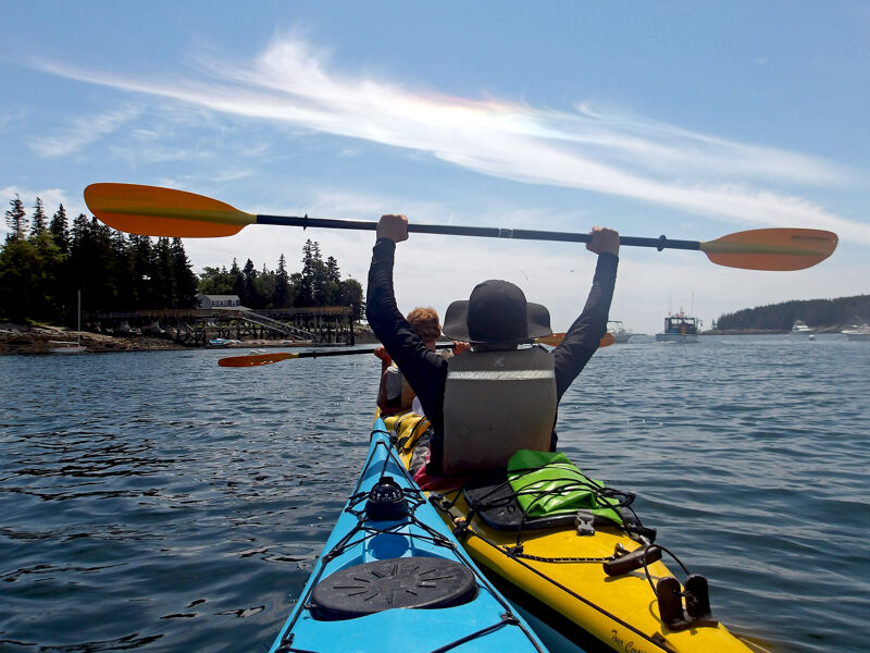 A group of people are kayaking on a sunny day. The person in the front kayak is holding their paddle up in the air with both hands. The kayaks are blue and yellow. The water is choppy and there are islands in the background.