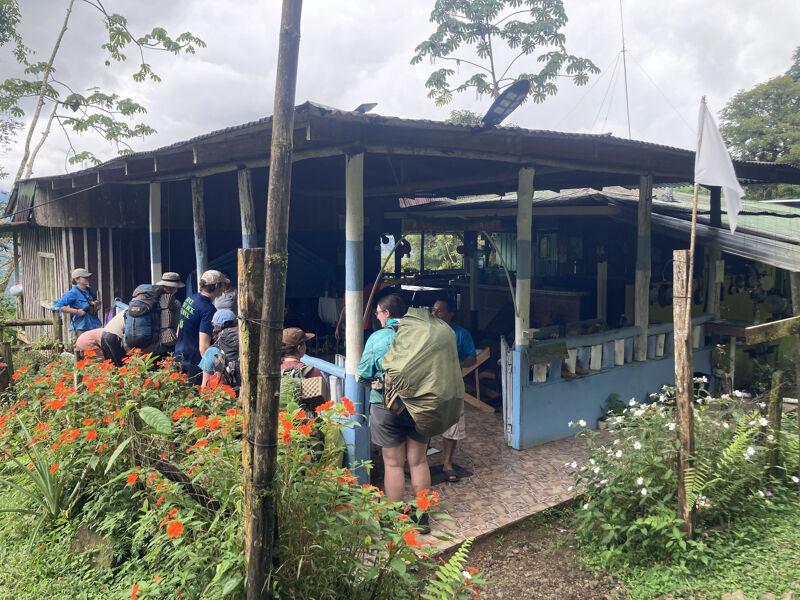 The image shows a group of people gathered near a building with a covered porch. The building appears to be in a rural or natural setting, with lush greenery and flowers in the foreground. A white flag is visible on the right side of the image. The people seem to be resting or preparing for an activity, possibly hiking or exploring the area.