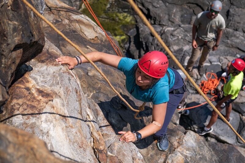 A woman in a blue shirt and red helmet is rock climbing. She is gripping the rock face with both hands and appears to be focused on her next move. A climbing rope is visible, secured above her. In the background, another person is standing on a rocky surface, and another person is climbing with a red helmet. The setting appears to be a natural rock formation, possibly a cliff or mountainside.