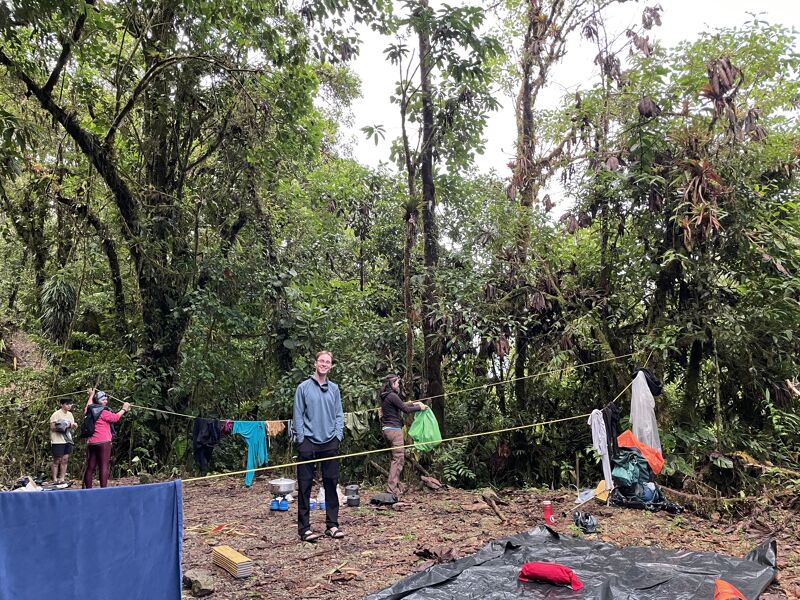 A man stands in a forest clearing, with a clothesline strung behind him holding various items. Other people are visible in the background, amidst lush greenery and tall trees. Camping gear is scattered around, suggesting an outdoor campsite. The scene conveys a sense of being immersed in nature, with the man as a central figure in this woodland setting.