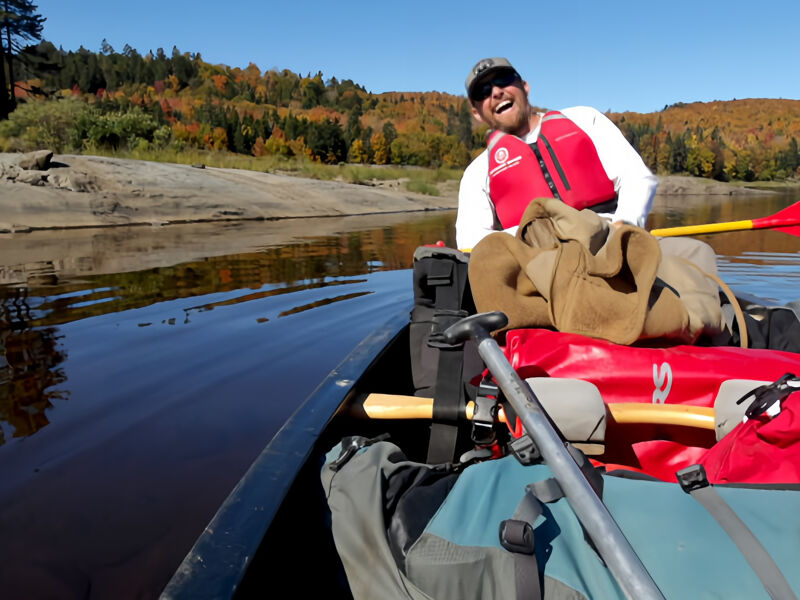A man wearing a red life jacket and sunglasses is sitting in a canoe on a calm river. The canoe is filled with gear, including bags and paddles. The river reflects the trees on the far bank, which are displaying autumn colors. The sky is blue and the sun is shining. The man is smiling and appears to be enjoying the scenery.