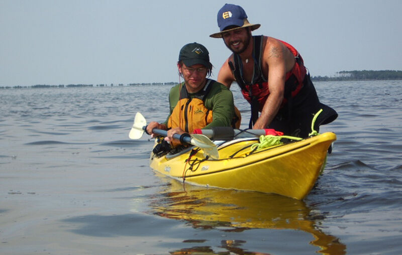 Two people are in a yellow kayak on the water. The person in the front is holding a paddle and wearing a life vest and a cap. The person in the back is wearing a hat, a life vest, and a tank top. The water is calm, and the sky is overcast. The kayak is reflecting in the water.