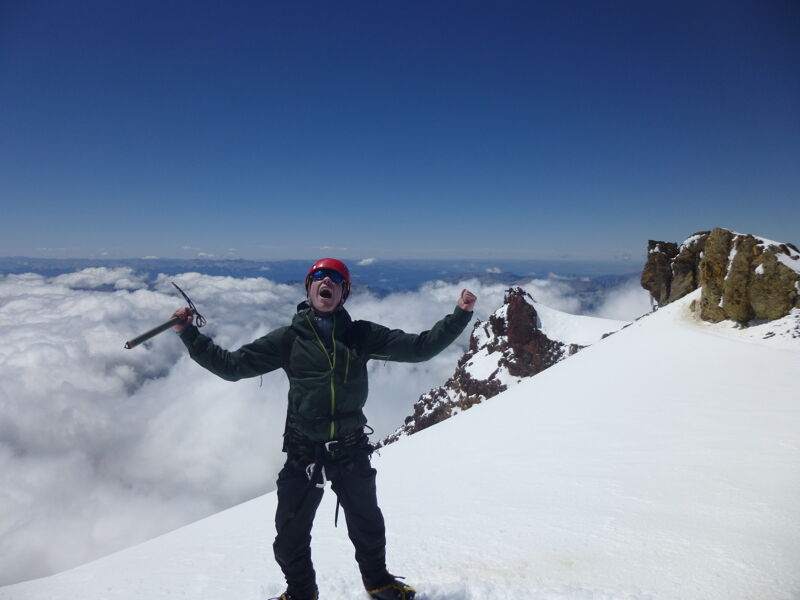 A person stands triumphantly on a snow-covered mountain peak under a clear blue sky. They are wearing dark clothing, a red headband, and sunglasses, and holding an ice axe in one hand. Clouds fill the valley below, creating a stunning backdrop. The surrounding landscape features rocky outcrops and pristine white snow, suggesting a challenging but rewarding climb.