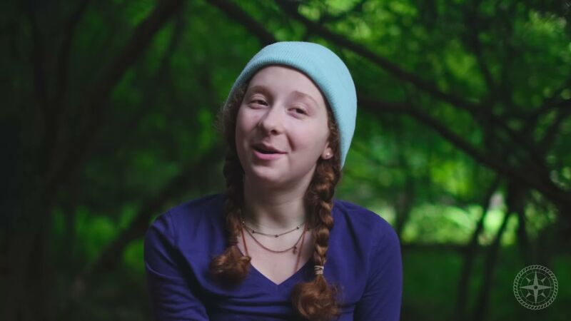 The image shows a young woman with reddish-brown hair styled in braids, wearing a blue beanie and a blue V-neck shirt. She is positioned in front of a lush green background of trees and foliage, suggesting an outdoor setting. The lighting is soft, highlighting her face and creating a natural, serene atmosphere. A logo is visible in the bottom right corner.