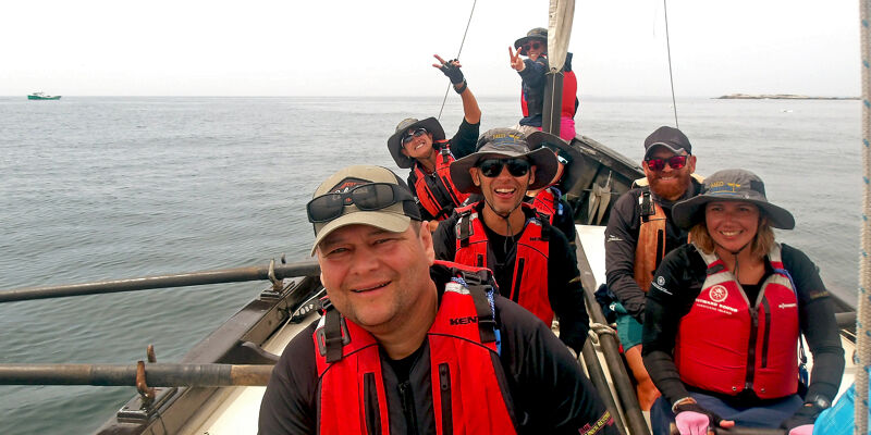 A group of people are on a boat, all wearing life jackets. They appear to be enjoying a sunny day out on the water. The boat is sailing on a calm sea, and the people are smiling and looking at the camera. There are at least six people visible in the image, and they are all dressed casually.
