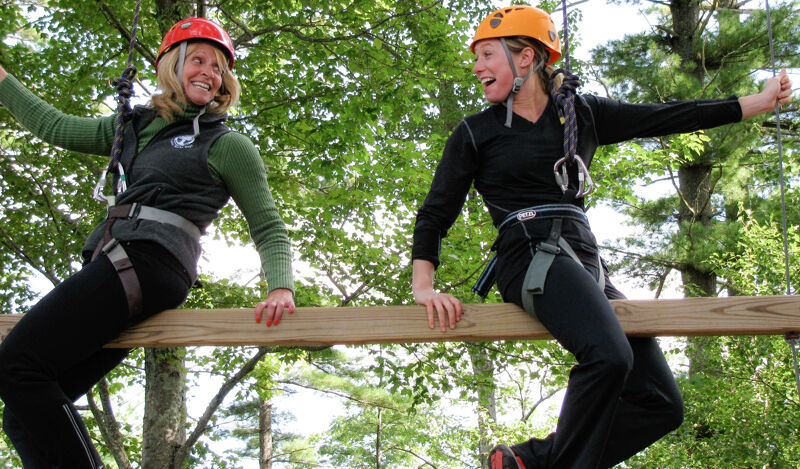 Two women are on a ropes course, balancing on a wooden beam. Both are wearing helmets and safety harnesses. The woman on the left has a red helmet and is smiling, while the woman on the right has an orange helmet and is looking at her companion with a smile. They are surrounded by trees and greenery.