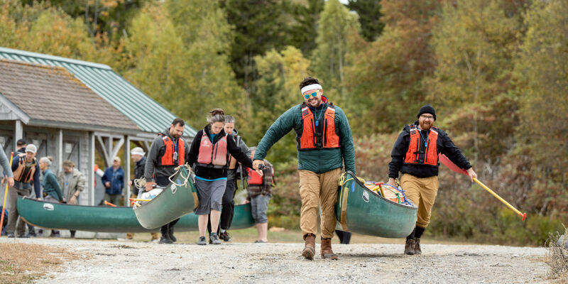 A group of people are carrying canoes near a building and a wooded area. Some are wearing life jackets. One person is using a paddle. The canoes are green. The people appear to be on a path or road. The building is light-colored with a green roof.