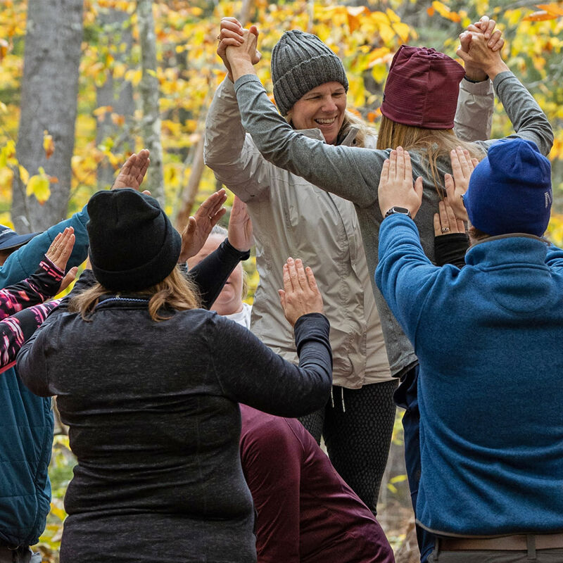 A group of people, mostly wearing beanies, stand in a circle outdoors, possibly in a forest or park. They are holding hands and raising their arms, suggesting a celebratory or team-building activity. The foliage in the background indicates it might be autumn, with yellow leaves visible. The people are dressed in casual, outdoor clothing, suitable for cooler weather.