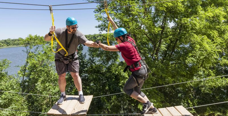 In the image, two people are on a ropes course, suspended high above the ground. The person on the left, wearing a gray shirt and blue helmet, is extending a hand to help the person on the right, who is wearing a red shirt and blue helmet. They are both connected to safety lines and are standing on wooden platforms. Lush green trees surround the course, and a body of water is visible in the background.