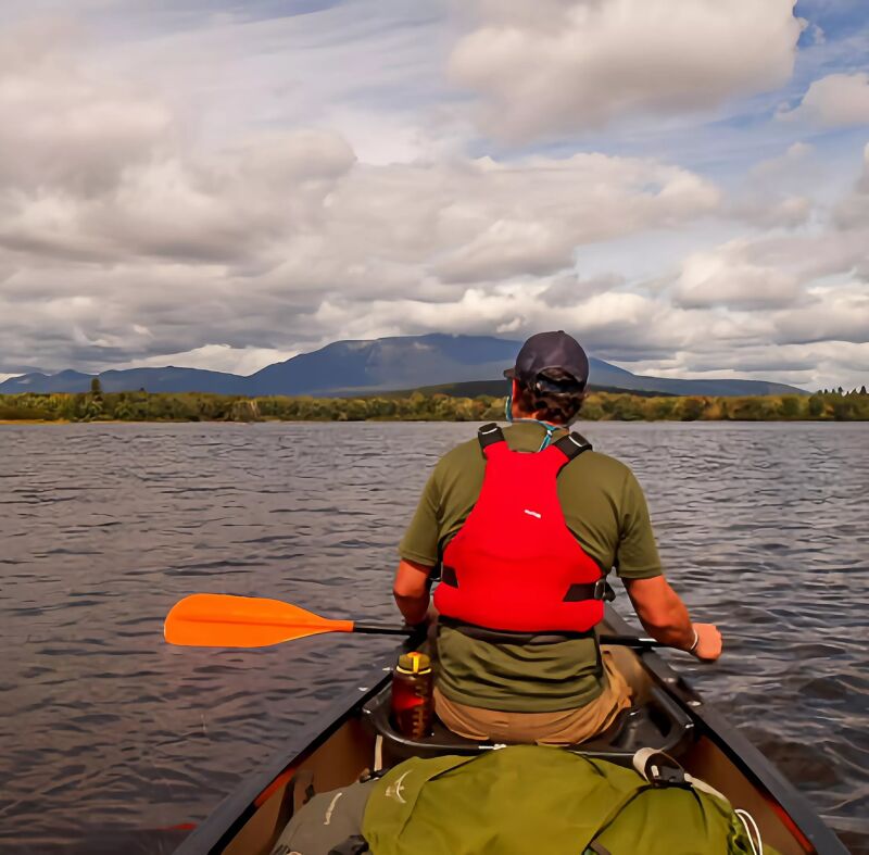 A person is paddling a canoe on a lake. The person is wearing a red life jacket and a hat. The canoe is in the foreground, and the lake and mountains are in the background. The sky is cloudy.