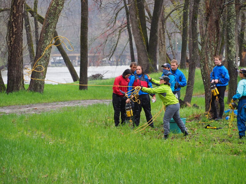 A group of people are participating in what appears to be a rescue training exercise near a body of water. They are using ropes and wearing casual clothing. The setting is a grassy area with trees, suggesting a park or natural environment. The weather seems overcast, and the overall scene conveys a sense of teamwork and preparedness.