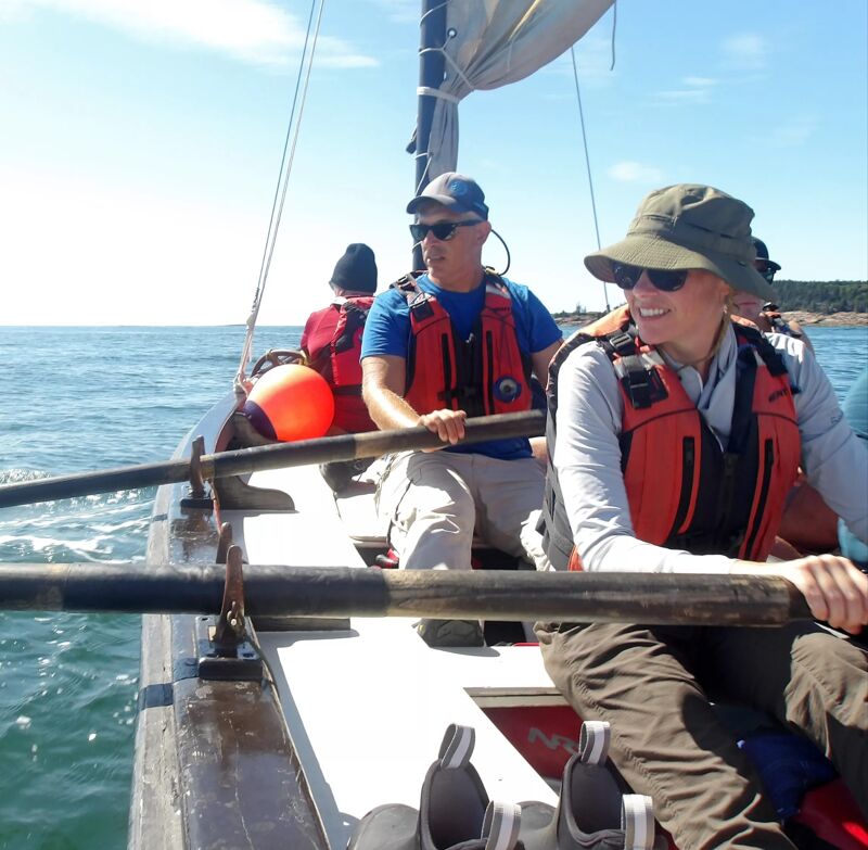 The image shows three people on a sailboat. The person in the foreground is wearing a hat and sunglasses and is holding an oar. Another person is next to them, also wearing sunglasses and a life jacket. A third person is in the background. They are sailing on a sunny day with blue skies and water.