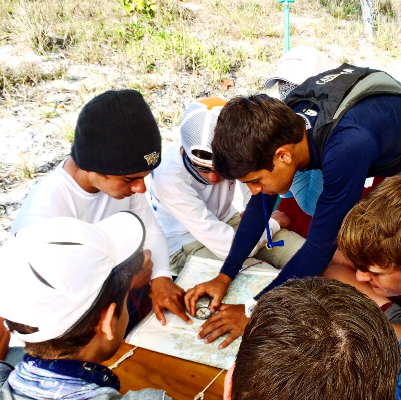 A group of people are gathered around a map, seemingly engaged in a navigation or orienteering activity. They appear to be outdoors, possibly in a wooded or natural area. One person is pointing at the map, while others are looking at it intently, suggesting they are working together to understand the map and plan their route.