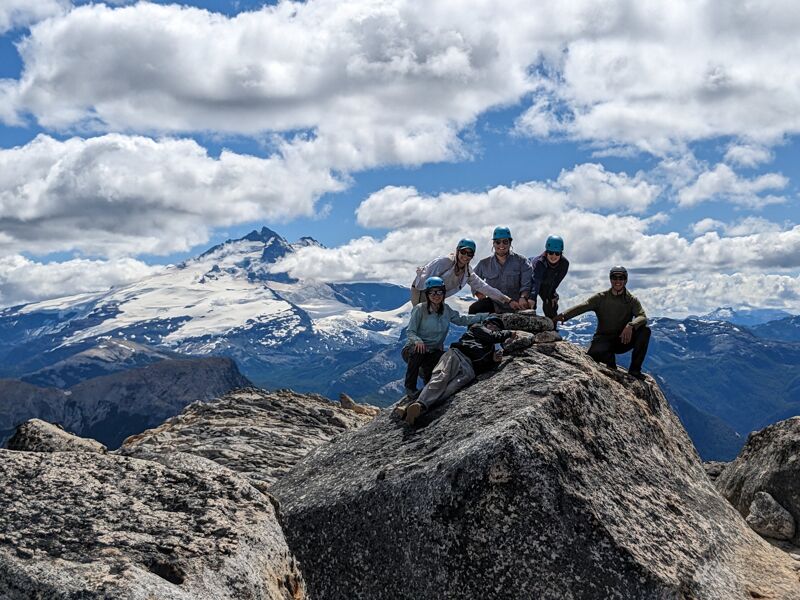 A group of five people are gathered on top of a rocky mountain peak, posing for a photo. They are surrounded by a vast landscape of mountains, some covered in snow, under a partly cloudy sky. The foreground is dominated by large boulders and rocky terrain, while the background features a panoramic view of the mountain range.
