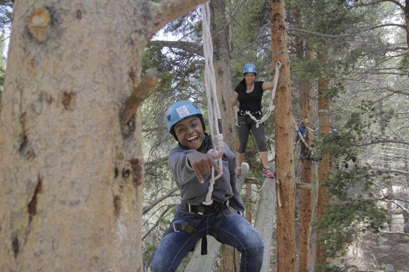 The image shows two women participating in a ropes course in a forest. The woman in the foreground is smiling and pointing towards the camera while holding onto a rope. She is wearing a helmet, a grey jacket, and jeans. The other woman is further back on the course, wearing a black shirt and helmet. Both women are secured with harnesses. The course is set up among tall trees.