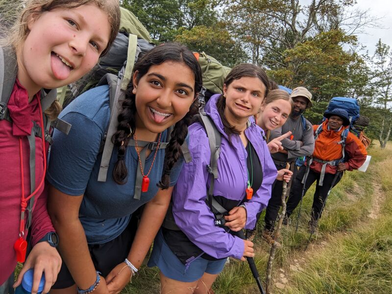 A group of seven young people are hiking outdoors. They are wearing backpacks and hiking clothes. They are smiling and appear to be enjoying their time. The background shows a grassy field and trees. The weather looks mild and overcast.