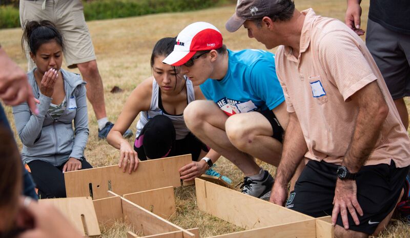 A group of people are gathered around a wooden structure on the grass. They appear to be collaborating or working together on a project, possibly involving construction or problem-solving. The individuals are dressed casually, and the setting seems to be outdoors, suggesting a team-building activity or a hands-on workshop.