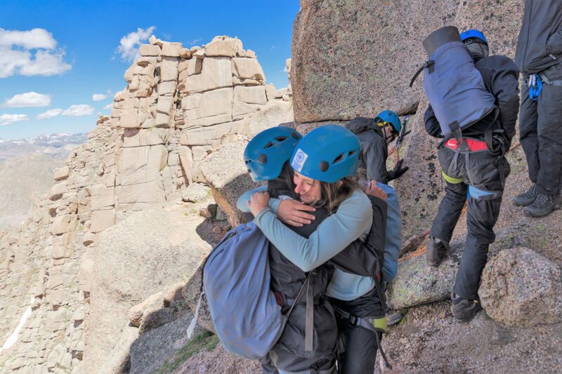 A group of people are climbing a rocky mountain. Two of them, wearing blue helmets and backpacks, are hugging each other. The landscape is rugged, with large rock formations and a distant view of the horizon. The sky is partly cloudy, suggesting a bright day for the climb.