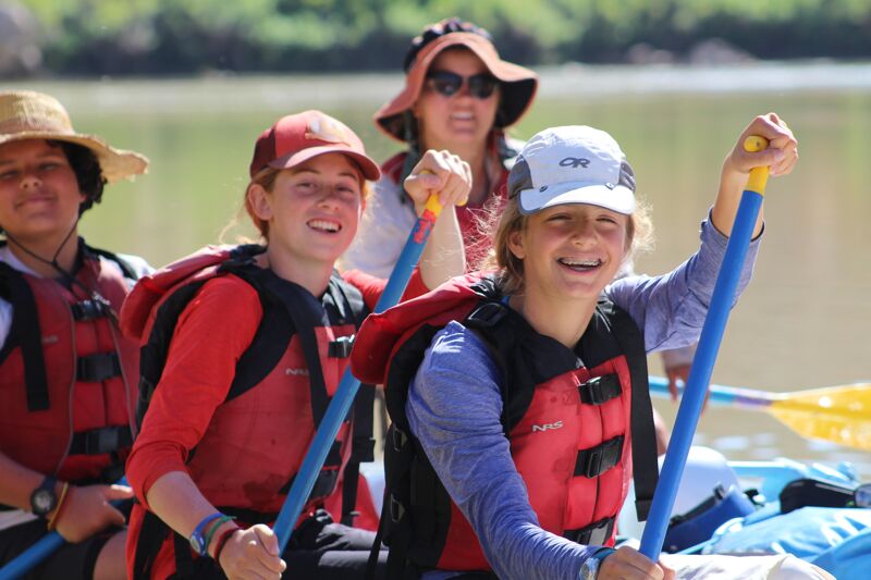 A group of four people are enjoying a sunny day on the water in kayaks. They are all wearing life jackets and hats, and they appear to be paddling together. The two people in the front are smiling and looking forward, while the two people in the back are partially visible. The background shows a calm river and green trees.