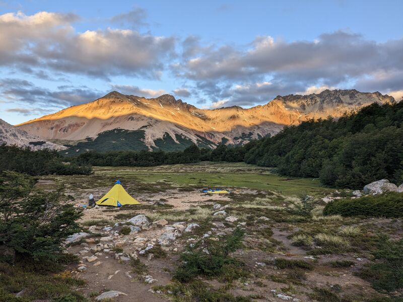 The image shows a scenic mountain landscape with a yellow tent pitched in a grassy field. The mountains in the background are illuminated by the warm light of the setting sun, casting long shadows across the landscape. The foreground features a mix of vegetation and rocky terrain, adding depth and texture to the scene. The sky is filled with clouds, creating a dramatic backdrop for the mountains.