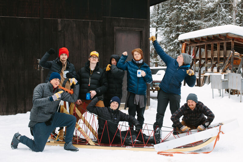 A group of people are gathered in a snowy outdoor setting, likely a winter camp or lodge. They are standing and sitting around a wooden sled, some holding snowballs, suggesting a playful atmosphere. The background features a dark wooden building and snow-covered trees, indicating a cold climate. The overall scene conveys a sense of camaraderie and winter fun.