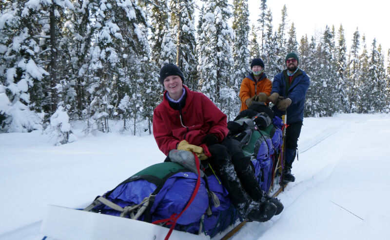 The image shows three people in a snowy, wooded area. They are on a sled that is loaded with gear. The person in the front is wearing a red jacket and a black hat and is smiling. The person in the middle is wearing an orange jacket. The person in the back is wearing a blue jacket and a black hat and has a beard. The trees in the background are covered in snow.