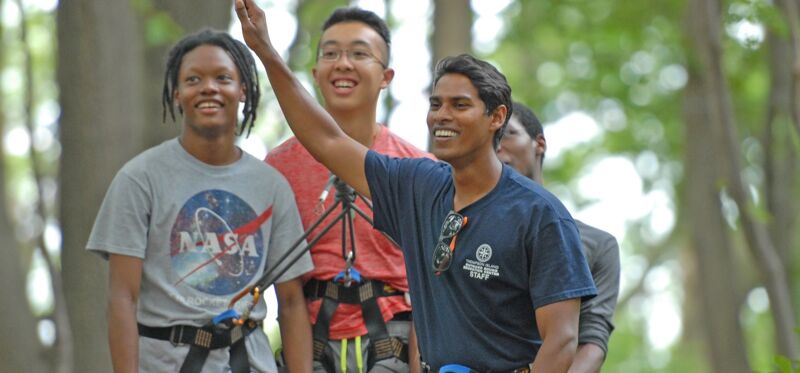 The image shows a group of young people outdoors, likely at an adventure park. They are wearing harnesses and appear to be engaged in an activity like zip-lining or climbing. The person in the center is raising their hand, and everyone seems happy and excited. The background is filled with trees, suggesting a natural setting.