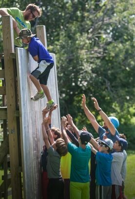 The image shows a group of people participating in an outdoor activity. A young person is climbing a slanted wall, assisted by others who are reaching up to help. The scene suggests teamwork and support, with individuals working together to overcome a challenge. The setting appears to be a camp or recreational area.