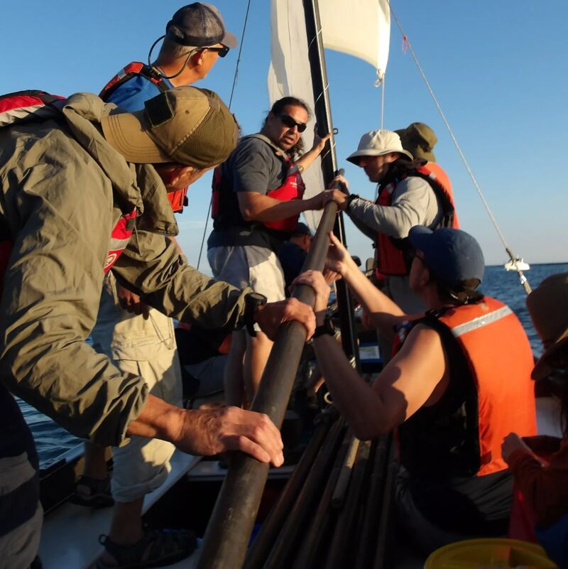 A group of people are on a sailboat, working together to raise or adjust the sail. They are wearing casual clothing and life vests, suggesting a recreational activity. The sky is clear, indicating fair weather for sailing. The focus is on teamwork and enjoying a day on the water.