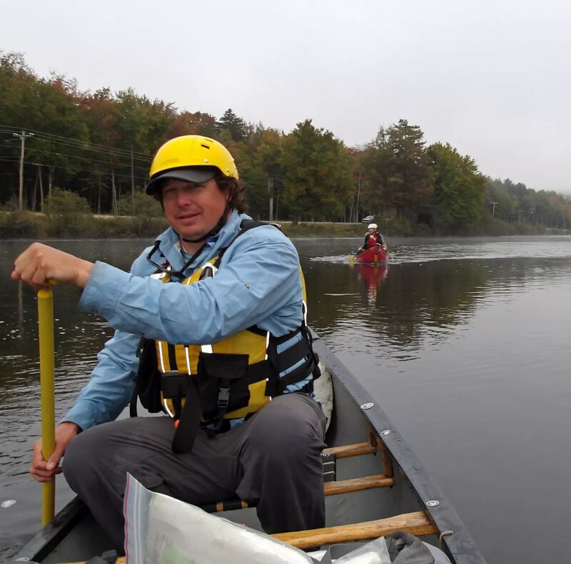 A man wearing a yellow helmet and life vest is paddling a canoe on a lake. He is holding a paddle in his right hand. Another person in a canoe is visible in the background. The water is calm, and the sky is overcast. Trees line the shore in the background.