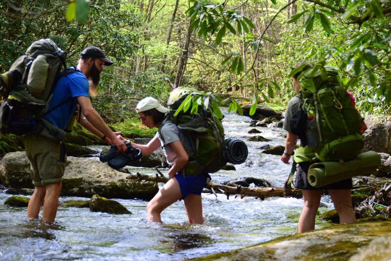 The image shows three people hiking through a shallow stream in a forest. They are all wearing large backpacks, indicating they are likely on a multi-day hike. The person in the front is handing their shoes to another person. The environment is lush and green, with trees lining the stream, suggesting a natural and possibly remote location.