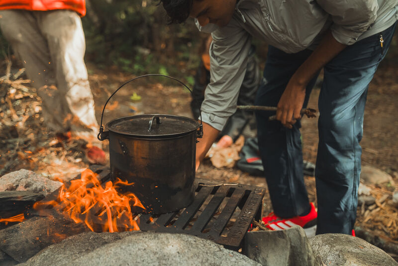 A person is tending to a campfire with a large black pot sitting on a grate over the flames. The individual is wearing jeans and red sneakers and appears to be adjusting the fire with a stick. Another person is partially visible in the background. The scene suggests they are cooking or boiling water over the open fire in an outdoor setting.