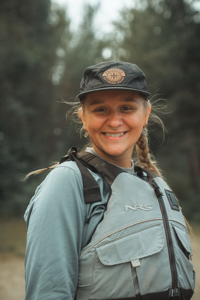A young woman with a braid smiles at the camera. She is wearing a black cap with a circular emblem, a light blue long-sleeved shirt, and a grey life vest with black straps. The background features blurred green trees, suggesting an outdoor setting, possibly near a river or lake. The lighting is soft and natural, highlighting her cheerful expression.