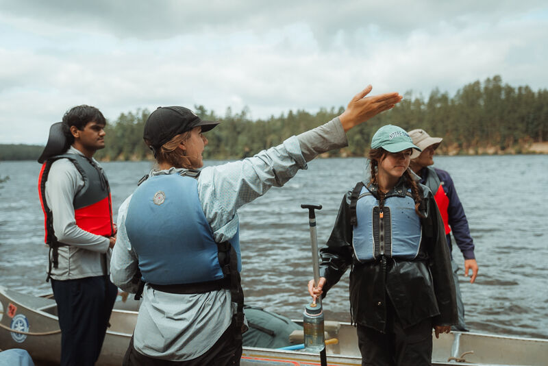 A group of four people are standing near a canoe on a cloudy day, seemingly preparing for a water activity. They are all wearing life jackets, suggesting a focus on safety. The person in the center is gesturing towards the water, possibly giving instructions or pointing out a direction. The background features a lake and a treeline, indicating a natural setting for their outing.