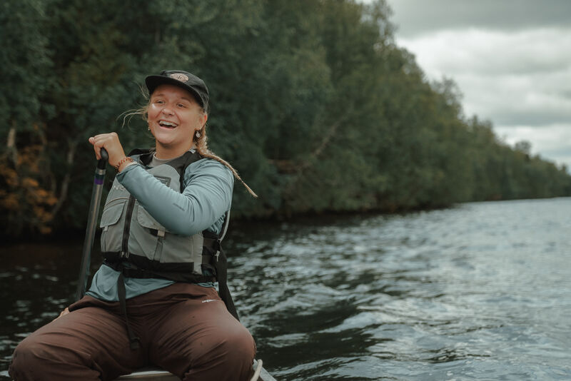 A woman wearing a life vest and a cap is paddling a canoe on a lake. She is smiling and appears to be enjoying the activity. The background features a lush green forest and a cloudy sky, suggesting a natural and possibly remote location. The water is calm with gentle ripples, indicating a peaceful day for canoeing.