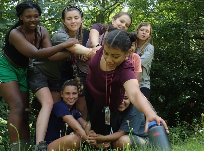 A group of young women are gathered outdoors, seemingly in a wooded area. They are interacting with each other, with some reaching out and holding onto one another. The woman in the center appears to be reaching down towards the ground. They seem to be engaged in a team-building activity or game. The overall atmosphere appears friendly and collaborative.
