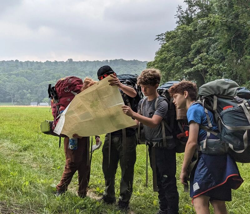 Four young hikers are gathered around a map in a grassy field, seemingly navigating their route. They are equipped with large backpacks, suggesting a multi-day trek. The weather appears overcast, with a hint of sunlight breaking through. The background features a line of trees and a distant hill, adding depth to the outdoor scene.