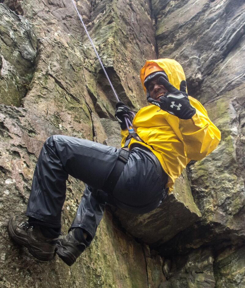 A person in a yellow raincoat and dark pants is rappelling down a rocky cliff face. They are wearing gloves and sturdy boots, and a rope is visible, guiding their descent. The person is giving a shaka sign with their right hand. The rock face is textured and appears damp, suggesting a recent rain or a naturally moist environment.