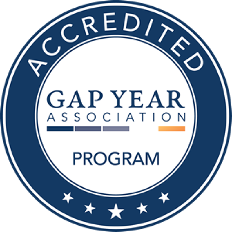 The image is a logo for an accredited Gap Year Association program. It features a circular design with the words "ACCREDITED" at the top and five stars at the bottom. The central part of the logo contains the text "GAP YEAR ASSOCIATION" with a horizontal line above and below it. The word "PROGRAM" is placed below the lower line. The logo has a blue and white color scheme.
