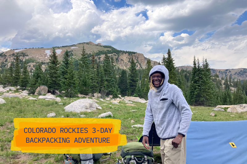 A man stands smiling in a mountainous landscape, likely in the Colorado Rockies, as suggested by the text overlay "Colorado Rockies 3-Day Backpacking Adventure." He is dressed in outdoor attire, with backpacking gear nearby, indicating he is on a hiking or camping trip. The background features a scenic view of mountains, trees, and a partly cloudy sky, contributing to the adventurous and outdoorsy feel of the image.