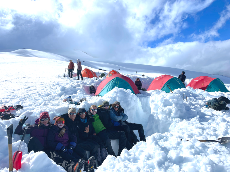 A group of people are huddled together in a snow trench, smiling and posing for a photo. Behind them, several colorful tents are pitched on the snowy landscape, suggesting a campsite in a mountainous or arctic environment. The sky is partly cloudy, with patches of blue visible.
