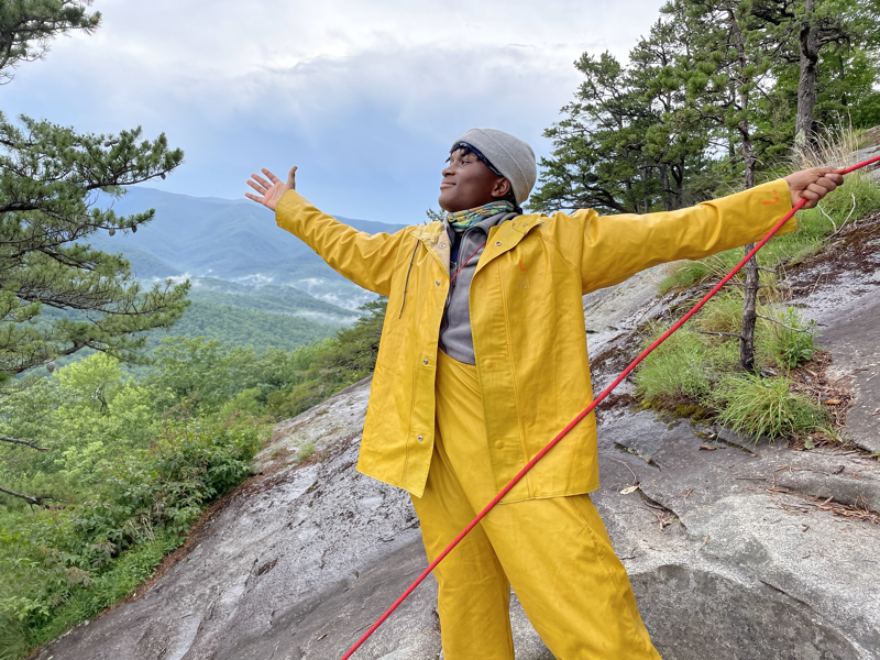 A person in a yellow raincoat and pants stands on a rocky outcrop, arms outstretched, with a scenic mountain view in the background. The sky is overcast, and trees surround the person. A red rope is visible, possibly indicating climbing or safety equipment. The person is wearing a head covering and appears to be enjoying the view and the moment.