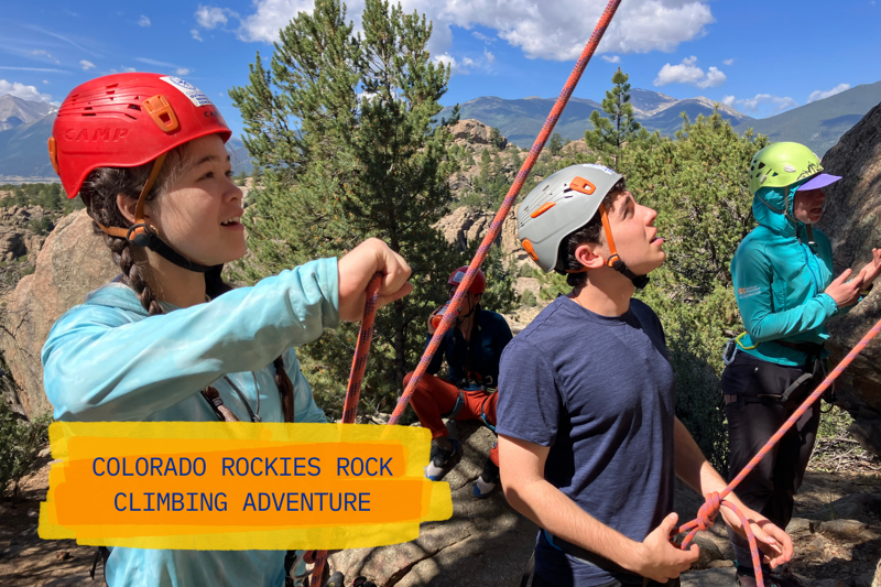 The image shows a group of people rock climbing in the Colorado Rockies. They are wearing helmets and harnesses, and ropes are visible, suggesting they are belaying each other. The climbers appear to be enjoying a sunny day outdoors, surrounded by trees and rocky terrain. The text overlay reads "Colorado Rockies Rock Climbing Adventure."