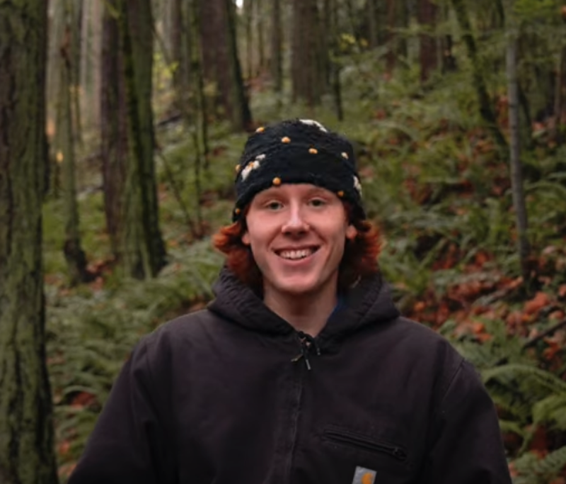 A young man with red hair and a black beanie with yellow flowers stands smiling in a forest. He is wearing a dark-colored Carhartt jacket. The background features lush green foliage and tall trees, creating a natural and woodsy setting. The man appears to be enjoying the outdoors, and the overall image has a cheerful and inviting feel.