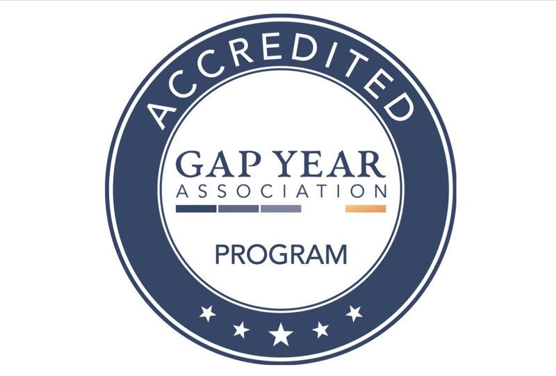 The image is a logo for the Gap Year Association Accreditation Program. It features a circular design with the words "ACCREDITED" at the top and "PROGRAM" at the bottom. The center of the logo contains the text "GAP YEAR ASSOCIATION" with a horizontal line design element. Five stars are spaced evenly along the bottom of the circle. The logo uses a combination of blue, white, and orange colors.
